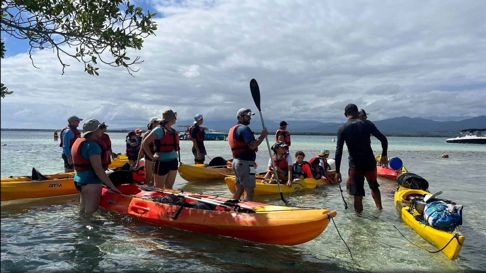 Un groupe de kayakistes en équipement de sécurité se prépare pour une excursion en mer en Guadeloupe. Ils sont à mi-chemin dans l'eau claire, à côté de leurs kayaks jaunes et rouges, écoutant un guide qui tient une pagaie. Le ciel est partiellement nuageux, et on peut voir des montagnes et un bateau de croisière en arrière-plan, offrant un contraste saisissant avec le paysage côtier tranquille.