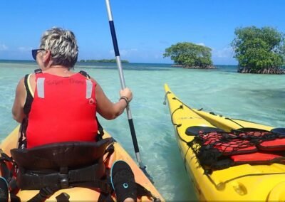 Une kayakiste en gilet rouge pagaye dans des eaux turquoises cristallines, approchant une petite île de mangrove. La tranquillité de l'expérience de kayak en pleine mer est mise en évidence par la clarté de l'eau et la sérénité du paysage.
